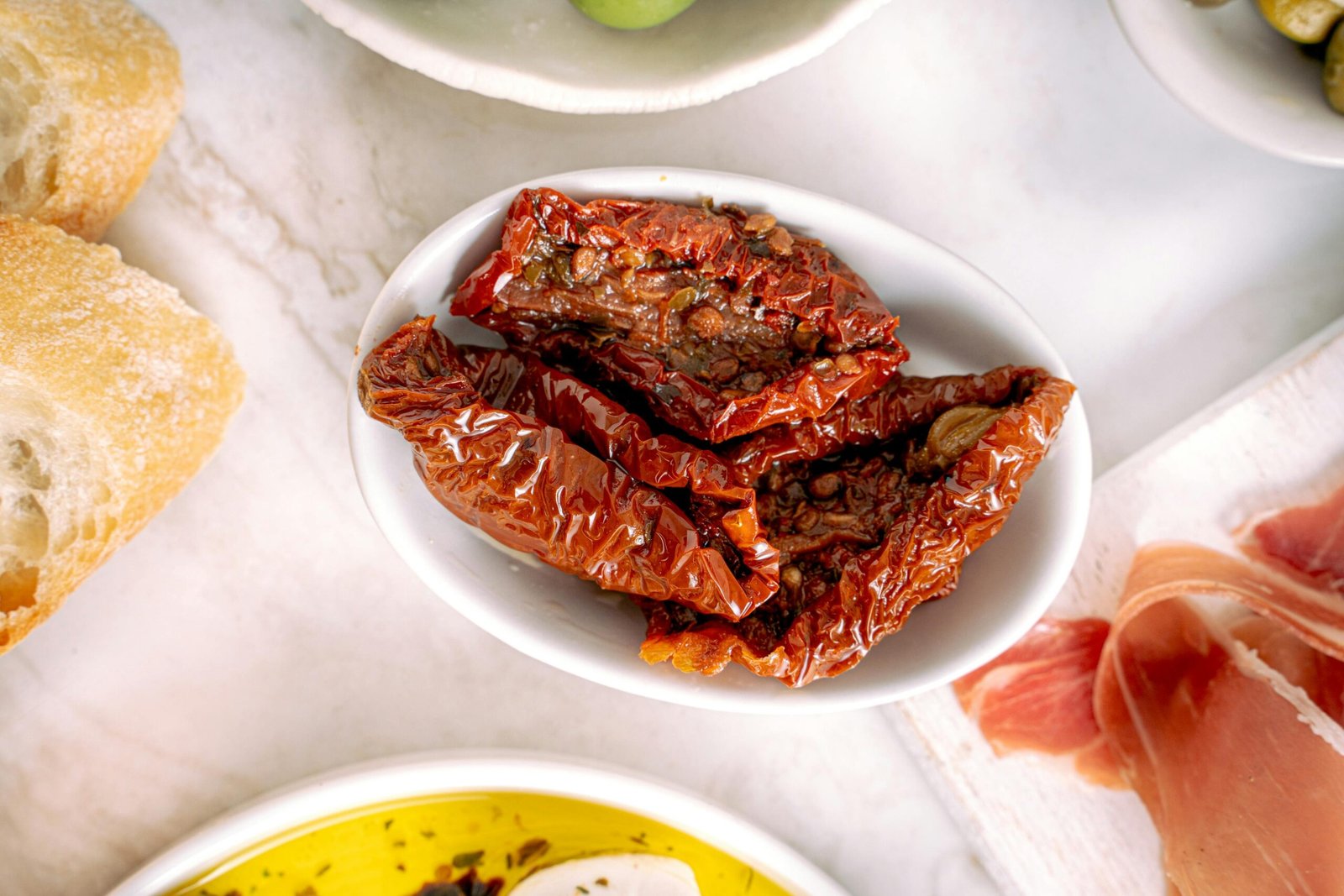 Content Image by Antoni Shkraba Studio Close-up of dried sun-dried tomatoes in a white bowl, with slices of fresh bread and olives in the background.
